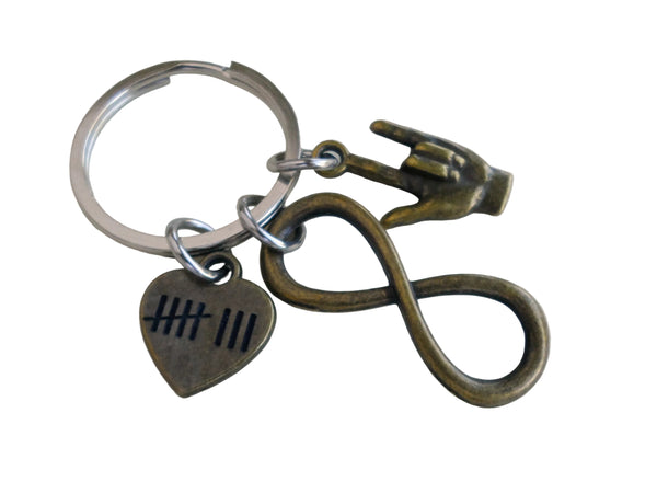 A metal keychain with a silver split ring, featuring an antique bronze infinity symbol charm, a heart-shaped charm with tally marks, and a small hand charm, shown on a plain light background.