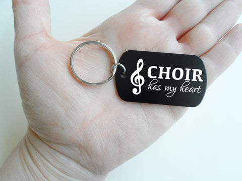 A black dog-tag style keychain with a silver metal key ring, featuring the white text 'CHOIR has my heart' and a white treble clef symbol, shown on a plain light background.